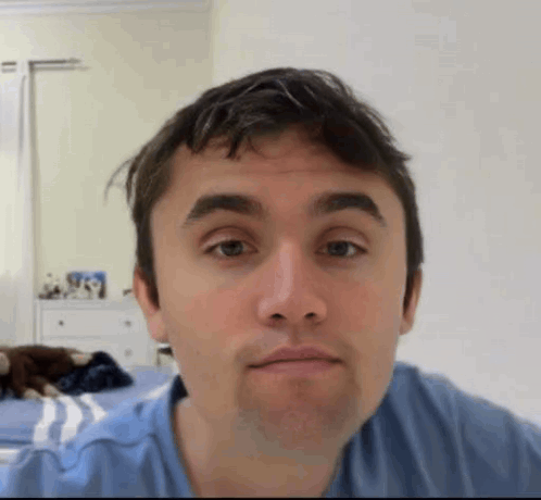 A Man In A Blue Shirt Is Making A Funny Face While Sitting On A Bed . GIF