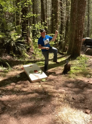 A Man In A Blue Shirt Is Playing A Game Of Cornhole In The Forest GIF