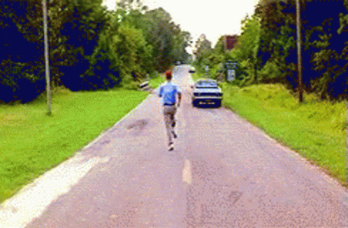 A Man In A Blue Shirt Is Running Down A Road With A Car In The Background GIF
