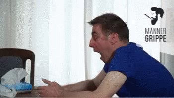 A Man In A Blue Shirt Is Sitting At A Table With His Mouth Open In Front Of A Sign That Says Manner Grippe GIF