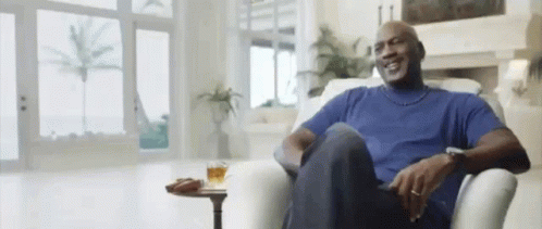 A Man In A Blue Shirt Is Sitting In A Chair In A Living Room With A Glass Of Whiskey On A Table . GIF