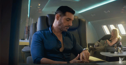 A Man In A Blue Shirt Is Sitting In A Chair Looking At His Watch GIF