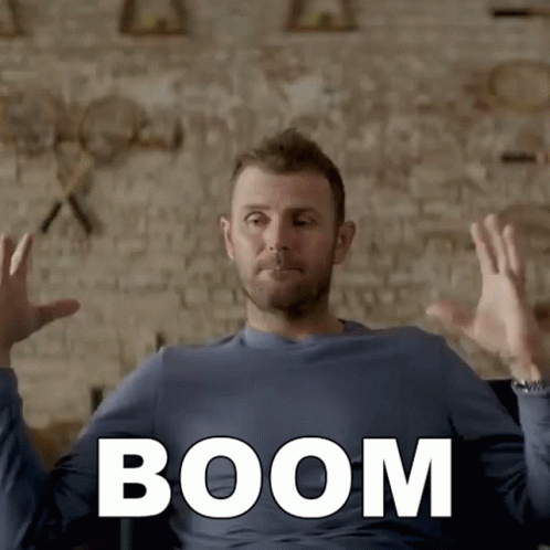 A Man In A Blue Shirt Is Sitting In Front Of A Brick Wall With The Word Boom Written On It GIF