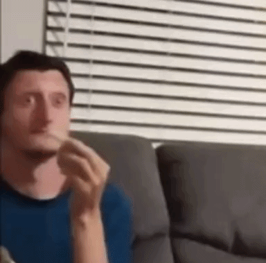 A Man In A Blue Shirt Is Sitting On A Couch Eating A Sandwich . GIF