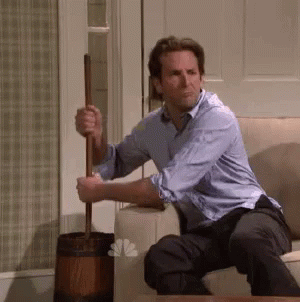 A Man In A Blue Shirt Is Sitting On A Couch Holding A Mop . GIF