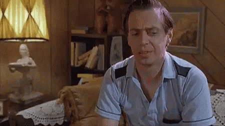 A Man In A Blue Shirt Is Sitting On A Couch In A Living Room With A Lamp In The Background . GIF