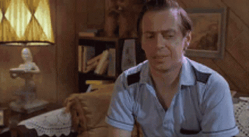A Man In A Blue Shirt Is Sitting On A Couch In A Living Room With A Lamp . GIF