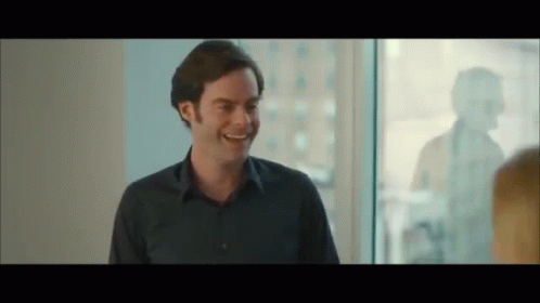 A Man In A Blue Shirt Is Smiling And Talking To Another Man In Front Of A Window . GIF