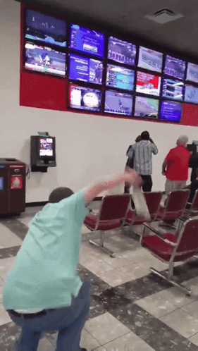 A Man In A Blue Shirt Is Squatting Down In Front Of A Wall Of Televisions Including One That Says ' Nfl ' GIF