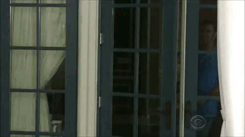 A Man In A Blue Shirt Is Standing In A Doorway Looking Out . GIF