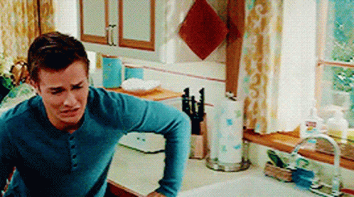 a man in a blue shirt is standing in a kitchen next to a sink and window .