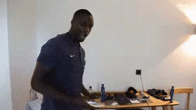 A Man In A Blue Shirt Is Standing In A Room Next To A Desk . GIF