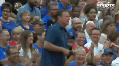 A Man In A Blue Shirt Is Standing In Front Of A Crowd Of People With Spectrum Sports Written On The Bottom Right GIF