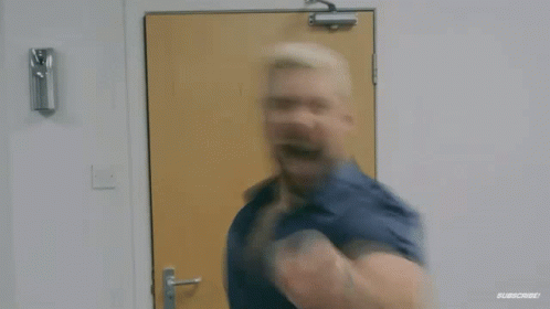 A Man In A Blue Shirt Is Standing In Front Of A Door And Making A Funny Face . GIF