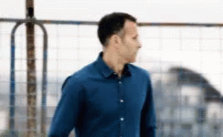 A Man In A Blue Shirt Is Standing In Front Of A Metal Fence . GIF
