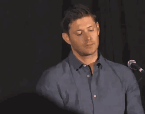 A Man In A Blue Shirt Is Standing In Front Of A Microphone With His Arm Raised GIF