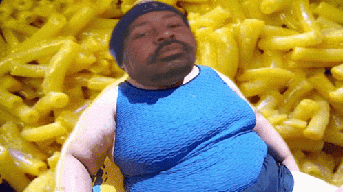 A Man In A Blue Shirt Is Standing In Front Of A Pile Of Macaroni And Cheese GIF