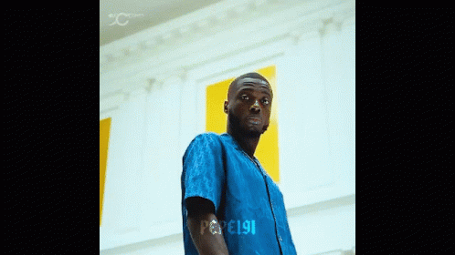 A Man In A Blue Shirt Is Standing In Front Of A Wall With The Number 191 On His Arm GIF