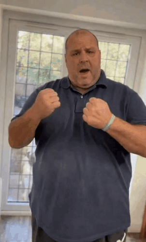 A Man In A Blue Shirt Is Standing In Front Of A Window With His Fists Up . GIF