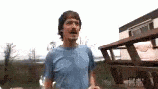 A Man In A Blue Shirt Is Standing In Front Of A Wooden Building . GIF