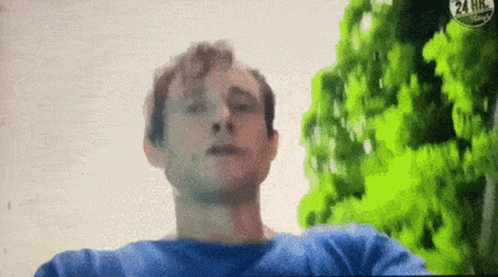 A Man In A Blue Shirt Is Standing In Front Of Trees And A 24 Hr Sign GIF