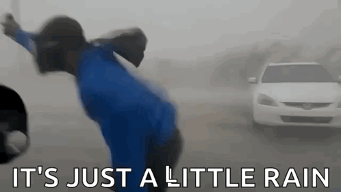 A Man In A Blue Shirt Is Standing In The Rain In Front Of A Car . GIF