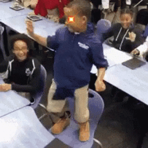A Man In A Blue Shirt Is Standing On A Chair In Front Of A Group Of People GIF