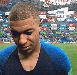 A Man In A Blue Shirt Is Standing On A Soccer Field With Ads For Qatar Coca Cola And Visa In The Background GIF