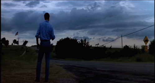 A Man In A Blue Shirt Is Standing On The Side Of A Road In Front Of A Road Sign GIF