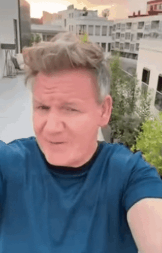 A Man In A Blue Shirt Is Taking A Selfie On A Rooftop With A City In The Background . GIF