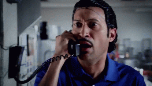 A Man In A Blue Shirt Is Talking On A Phone With A Surprised Look On His Face GIF