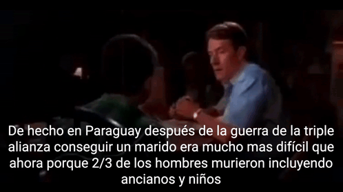 a man in a blue shirt is talking to another man in a dark room in spanish