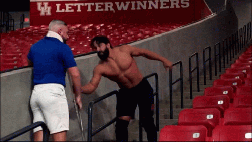 A Man In A Blue Shirt Is Talking To Another Man In Front Of A Sign That Says Letterwinners On It GIF