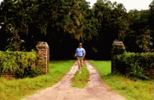 A Man In A Blue Shirt Is Walking Down A Dirt Road . GIF