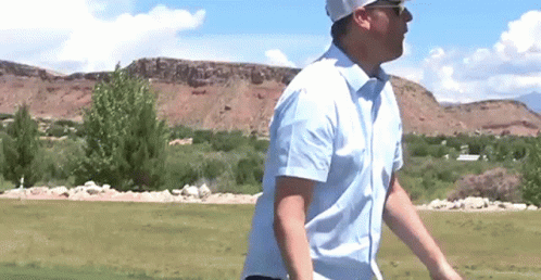 A Man In A Blue Shirt Is Walking On A Golf Course . GIF