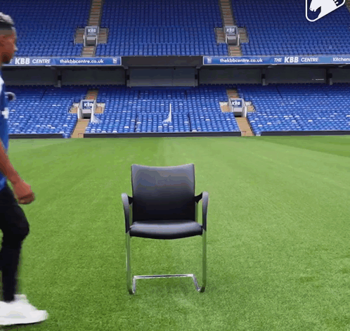 a man in a blue shirt is walking on a soccer field with a chair in the foreground that says kbb centre
