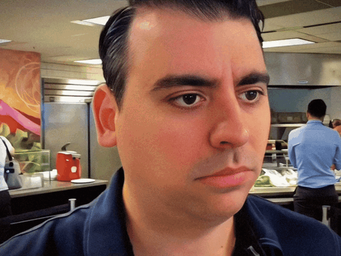 A Man In A Blue Shirt Looks At The Camera With A Red Ketchup Dispenser In The Background GIF