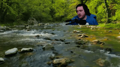 A Man In A Blue Shirt Sits In Front Of A Stream GIF
