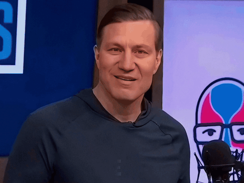 A Man In A Blue Shirt Stands In Front Of A Microphone In Front Of A Sign That Says S GIF
