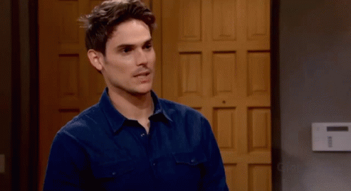 A Man In A Blue Shirt Stands In Front Of A Wooden Door GIF