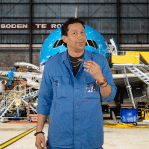 A Man In A Blue Shirt Stands In Front Of An Airplane In A Hangar With Boden Te Ro Written On The Wall Behind Him GIF