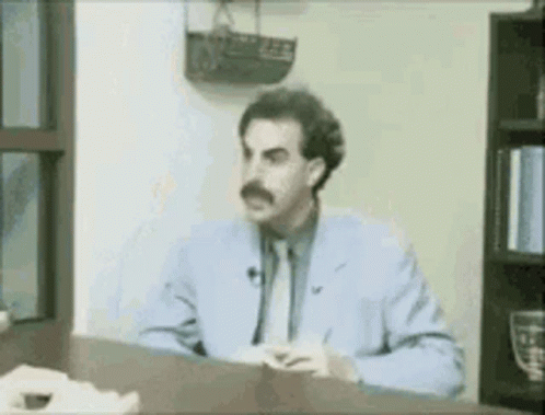 A Man In A Blue Suit And Tie Is Sitting At A Desk In Front Of A Window . GIF