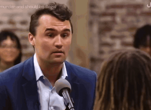 A Man In A Blue Suit Is Talking Into A Microphone While A Woman Looks On . GIF
