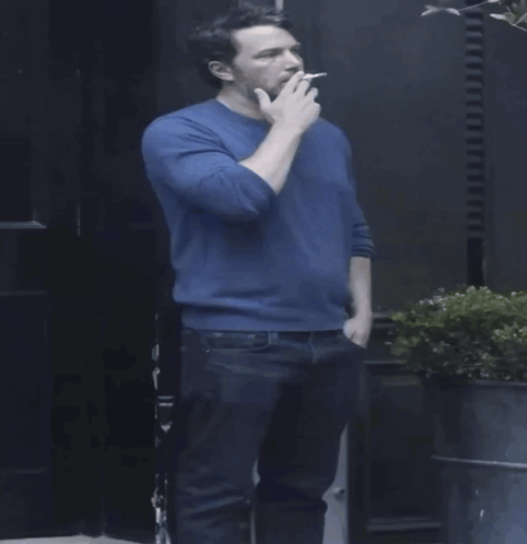 A Man In A Blue Sweater Holds A Cigarette GIF