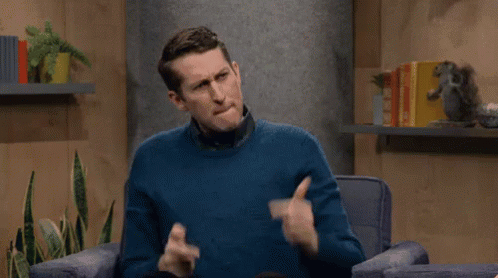 A Man In A Blue Sweater Is Sitting In A Chair Making A Gesture With His Hands . GIF