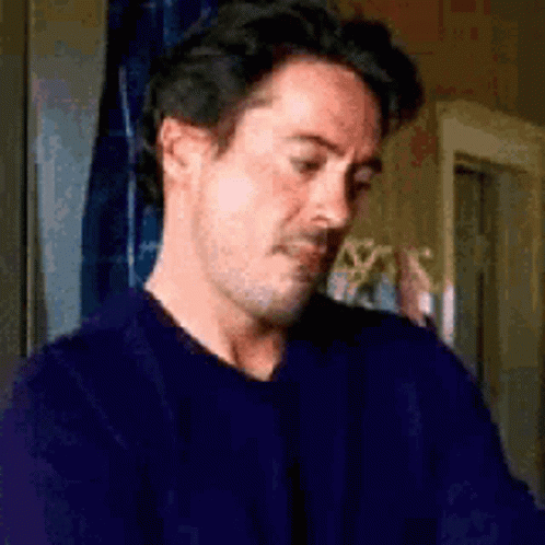 A Man In A Blue Sweater Is Sitting In Front Of A Window And Looking Down . GIF