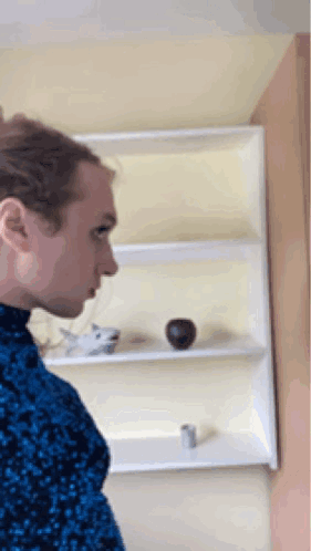 A Man In A Blue Sweater Is Standing In Front Of A Shelf With A Vase On It GIF