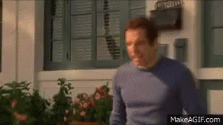 A Man In A Blue Sweater Is Standing In Front Of A Window With Shutters On It . GIF