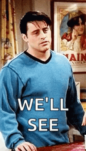 A Man In A Blue Sweater Is Standing Next To A Table With The Words `` We 'Ll See '' Written On It . GIF
