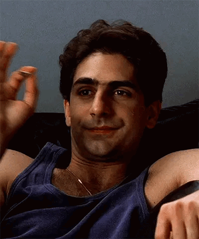 A Man In A Blue Tank Top Is Sitting On A Couch With His Arms Outstretched And Smiling . GIF